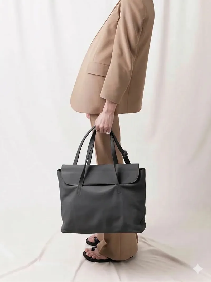 Model in a beige suit holding a dark leather tote against a cream backdrop